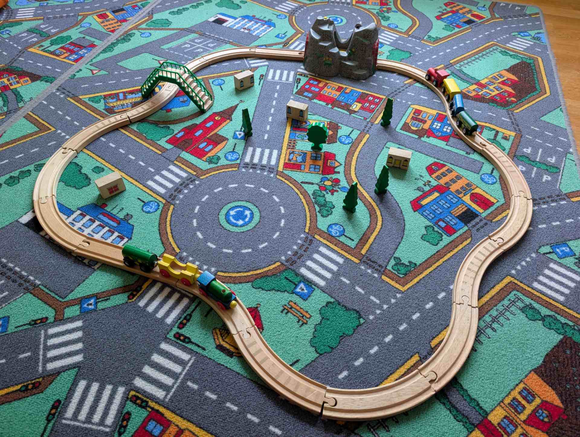 Eine kreativ aufgebaute Holzeisenbahn mit Brücke, Bergtunnel und kleinen Bäumen auf dem Spielteppich in unserer Tagespflege.