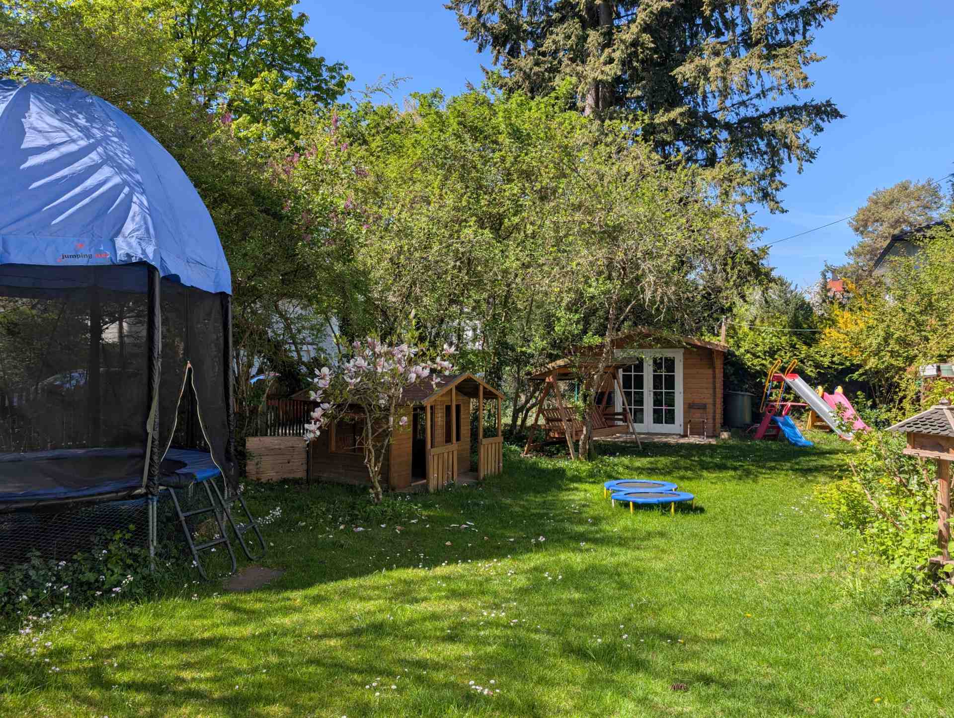 Unser weitläufiger Kita-Garten zum Toben, ausgestattet mit einem großen Trampolin, einem Holzspielhaus und viel grüner Rasenfläche unter Bäumen.