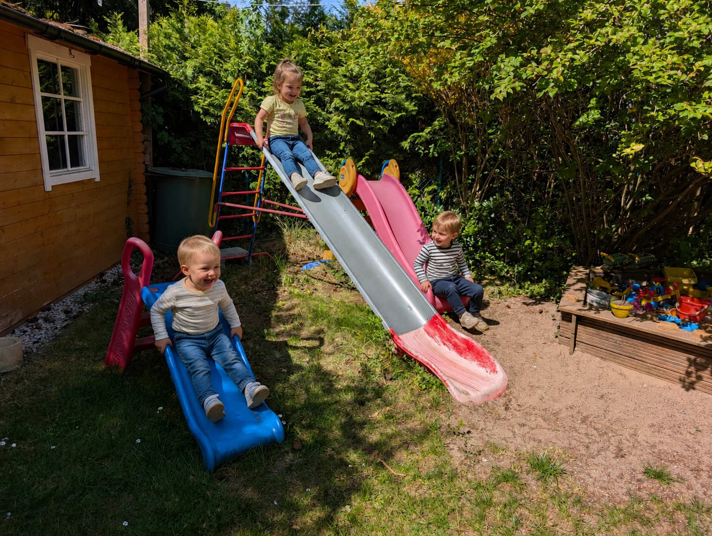 Drei lachende Kinder rutschen begeistert auf verschiedenen bunten Kinderrutschen in unserem großen Garten neben dem Sandkasten.