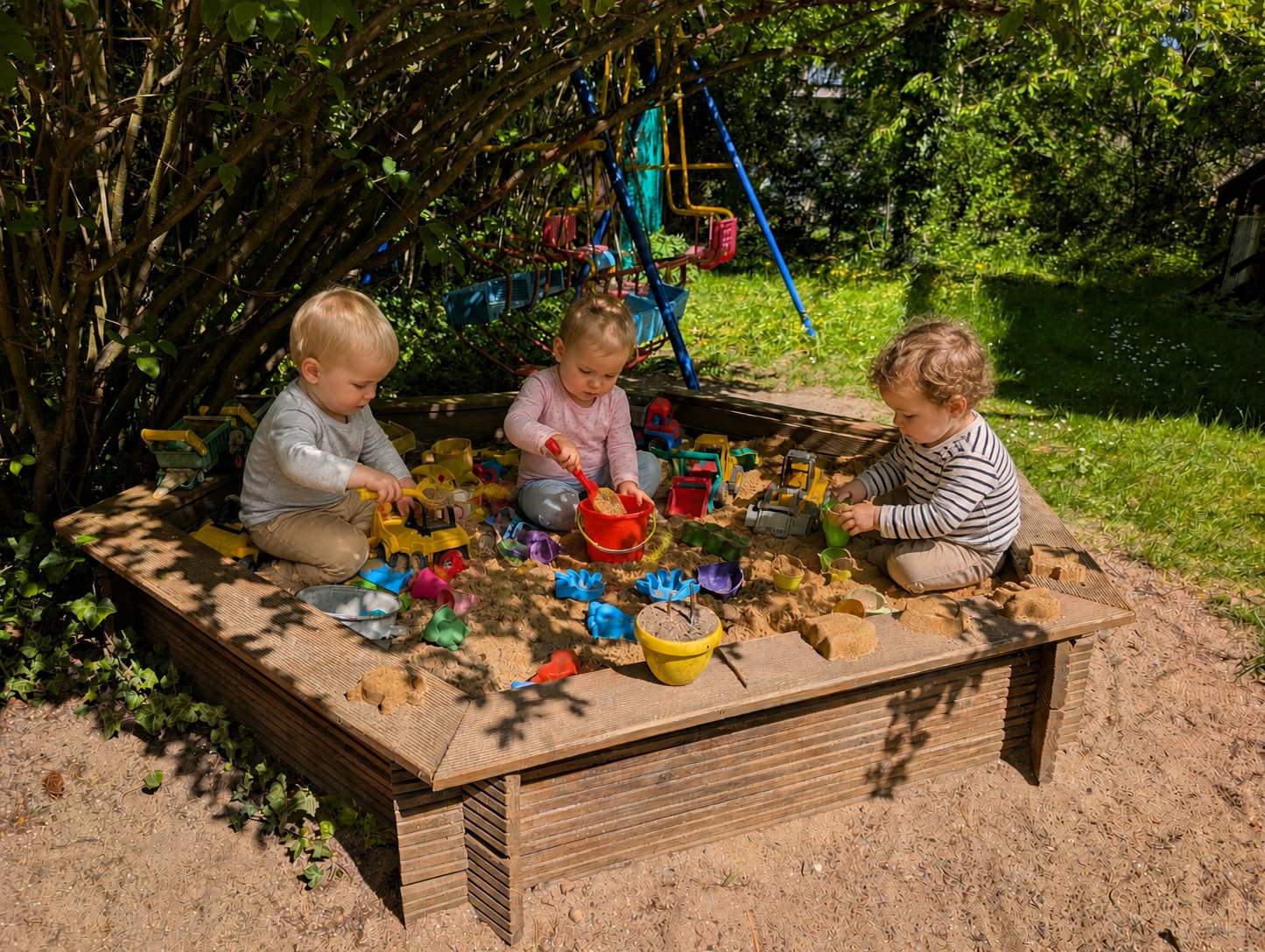 Drei Kleinkinder spielen konzentriert zusammen mit Eimern, Schaufeln und Spielzeugbaggern im schattigen Sandkasten unserer Tagespflege.