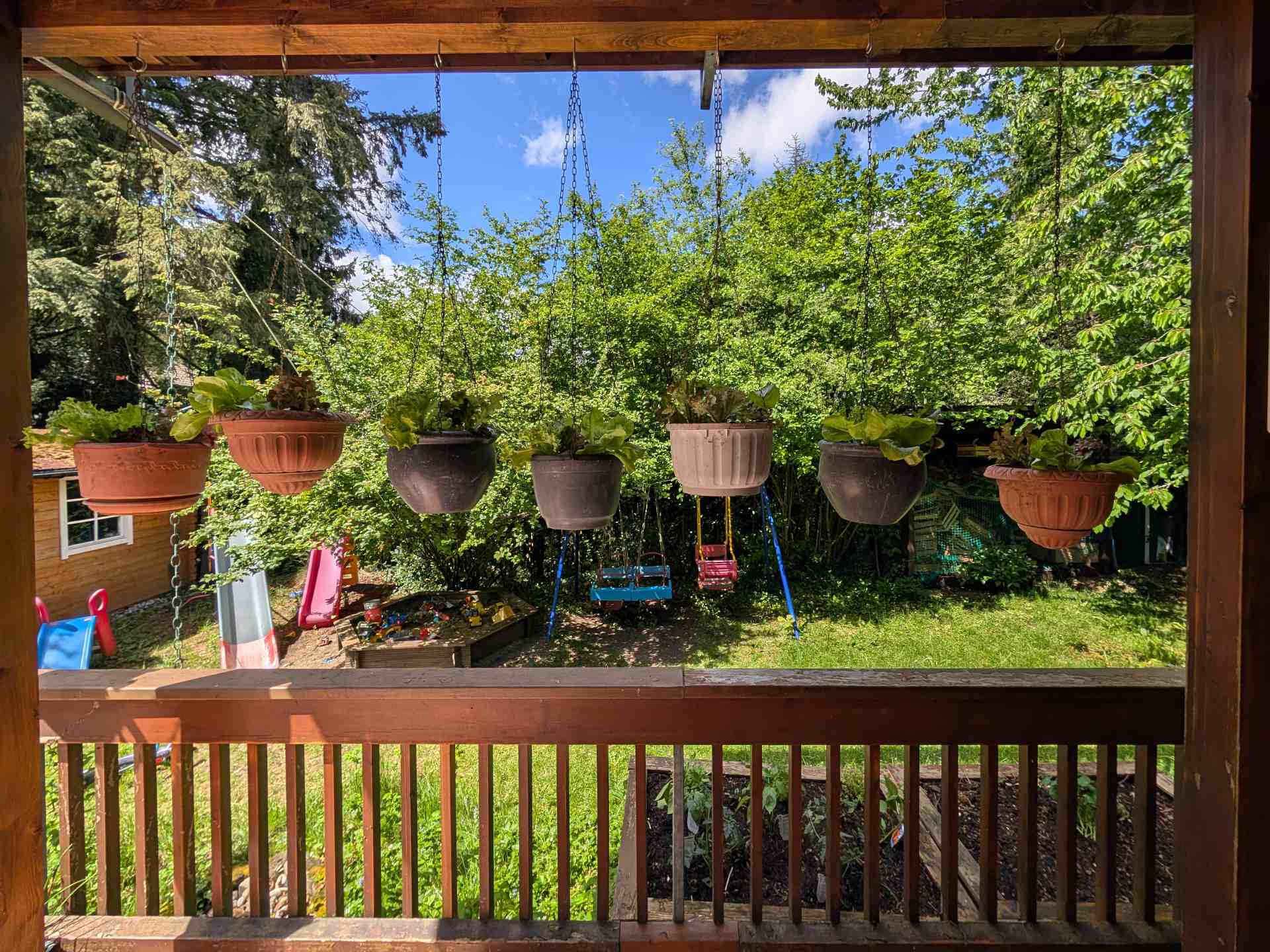 Blick von der überdachten Holzterrasse in den grünen Garten, geschmückt mit hängenden Pflanztöpfen. Im Hintergrund warten eine Kinderschaukel und der Sandkasten.
