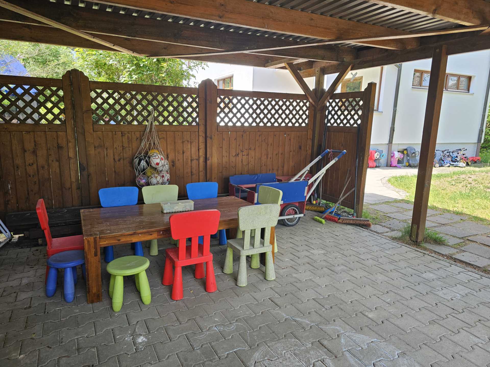 Überdachter, schattiger Außenbereich mit einem Holztisch und bunten Kinderstühlen, ideal für eine kleine Pause im Freien.